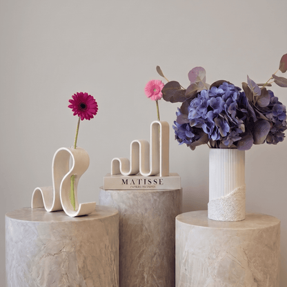 Vaze decorative Vernisaj alb de la Antiform, forme ondulate sculpturale cu gerbera roz și hortensii mov, pe piedestale de marmură