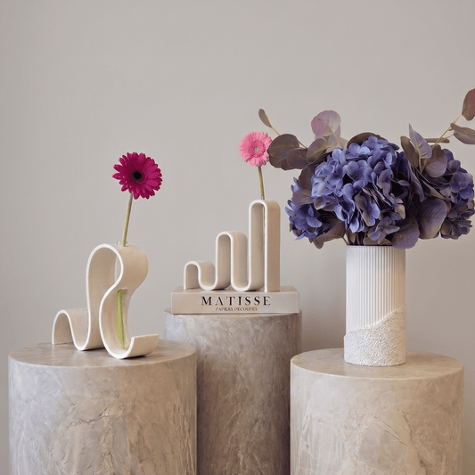 Vaze decorative Vernisaj alb de la Antiform, forme ondulate sculpturale cu gerbera roz și hortensii mov, pe piedestale de marmură